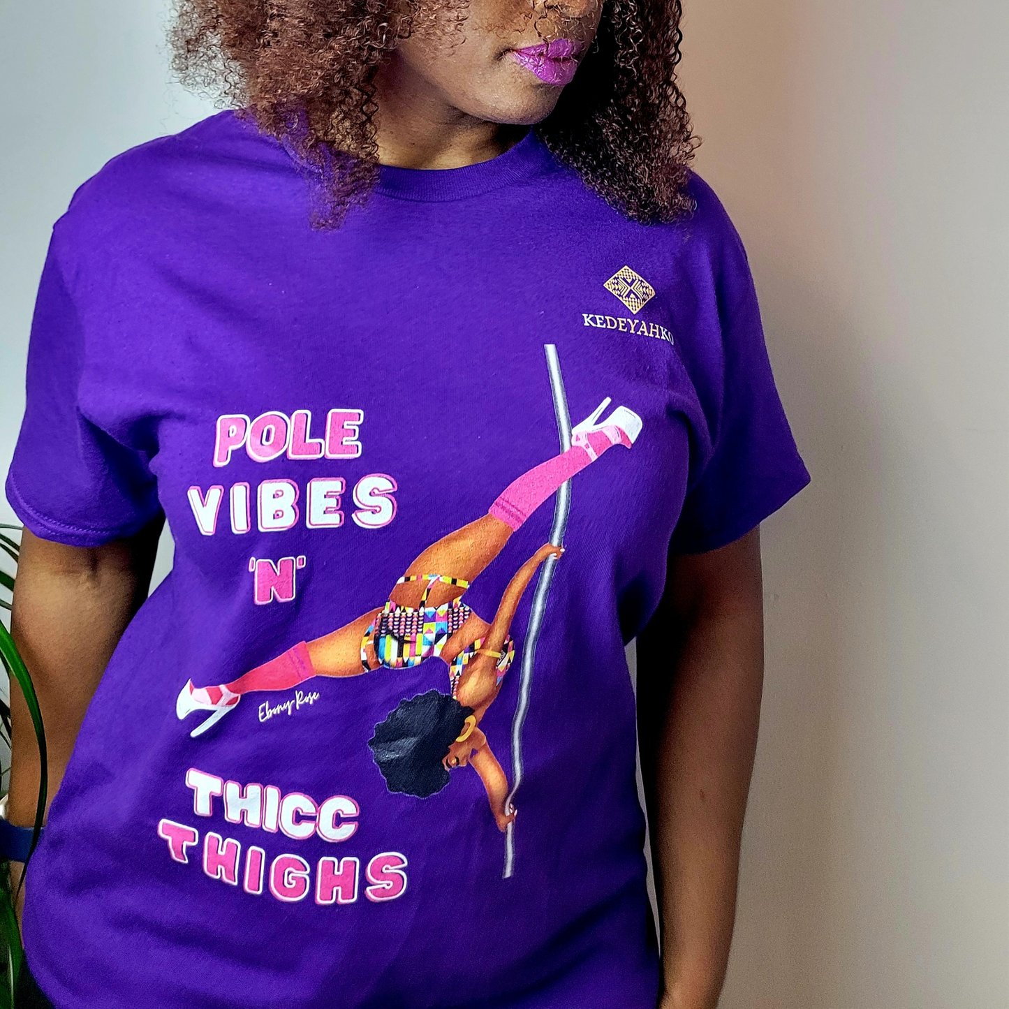 Purple unisex t-shirt  featuring a black pole dancer wearing bright African inspired Afrocentric garter pole shorts and sports top. Bright pink knee socks and clear platform heels in an extended butterfly pole pose with slogan affirmation pole vibes n thicc vibes