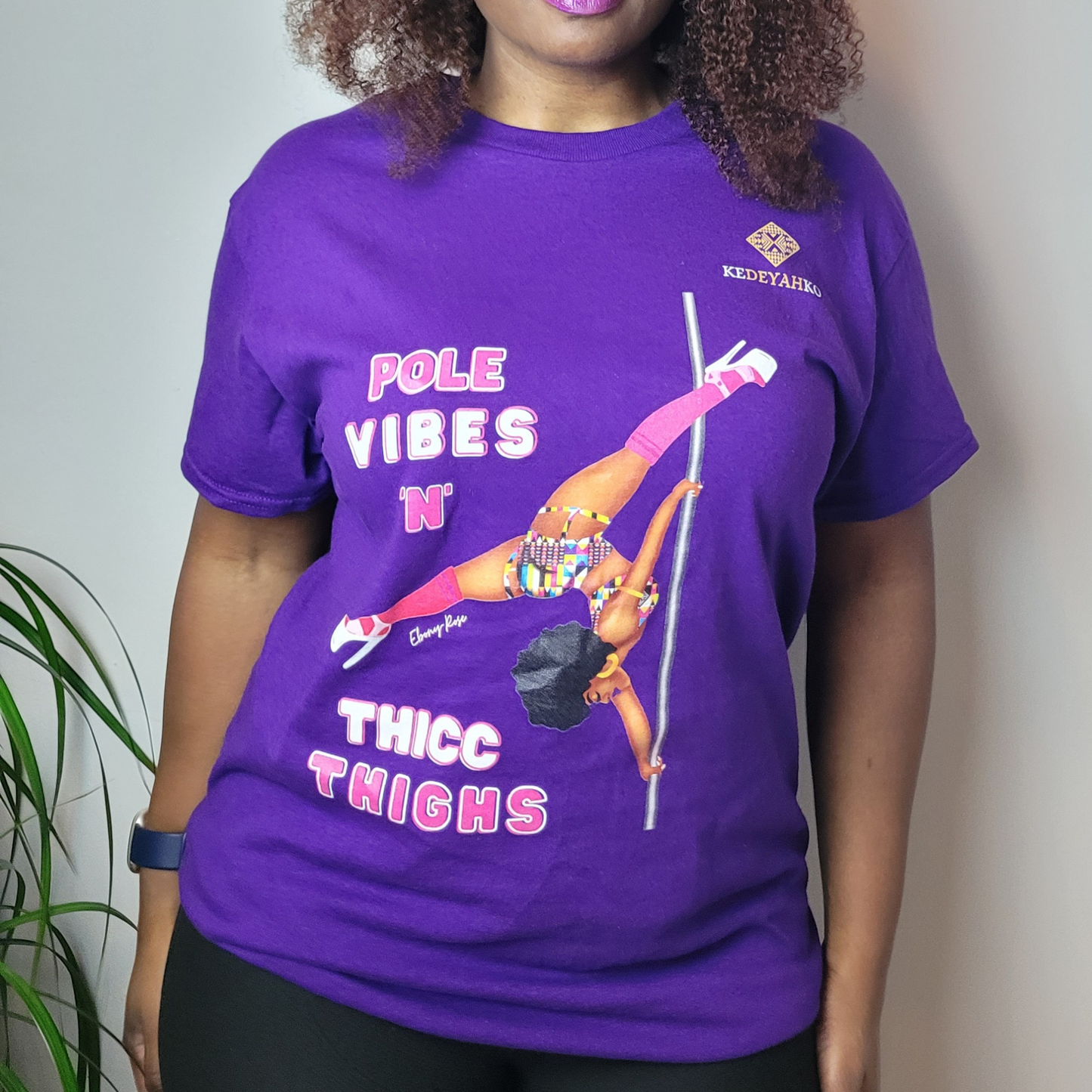 Purple unisex t-shirt  featuring a black pole dancer wearing bright African inspired Afrocentric garter pole shorts and sports top. Bright pink knee socks and clear platform heels in an extended butterfly pole pose with slogan affirmation pole vibes n thicc vibes