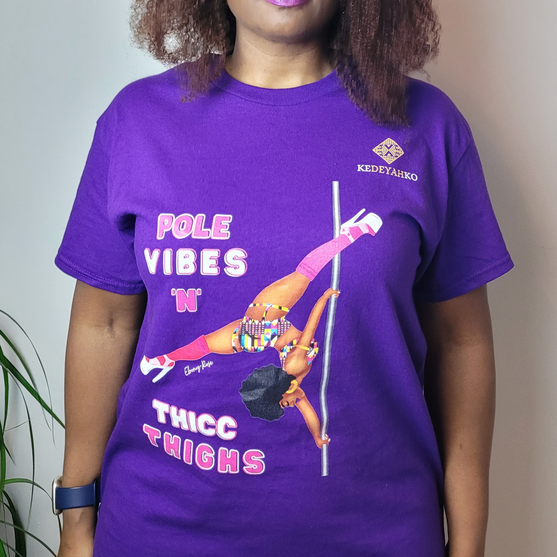 Purple unisex t-shirt  featuring a black pole dancer wearing bright African inspired Afrocentric garter pole shorts and sports top. Bright pink knee socks and clear platform heels in an extended butterfly pole pose with slogan affirmation pole vibes n thicc vibes