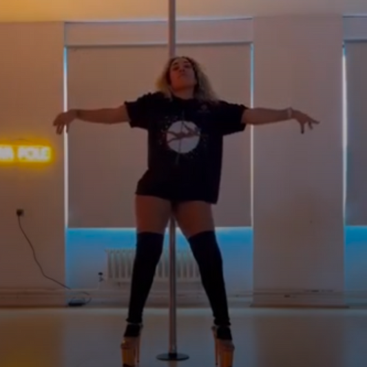 Mixed-race pole dancer with blonde hair performing a pose in a pole studio, wearing a black Kedeyahko T-shirt featuring the image of a Black pole dancer doing the Superman pole pose, knee-high socks, and heels